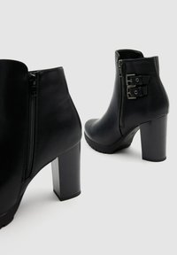 Botas de piel negra hasta el tobillo con un alto tacón de bloque. Cuentan con una cremallera lateral y acentos de doble hebilla en el lado exterior. Textura suave.