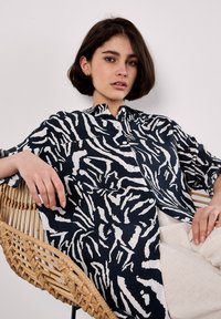 Una giovane donna con capelli corti e scuri è seduta su una sedia intrecciata, indossando una camicia a righe blu e bianca, con uno sfondo neutro.