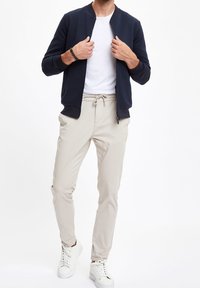 Granatowa kurtka bomber z teksturowanej tkaniny, biała koszulka, beżowe spodnie z sznurkiem oraz białe trampki, przedstawiające casualowy wielowarstwowy strój.