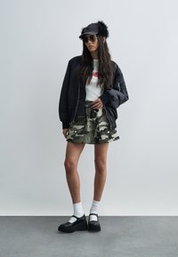 Γυναίκα που φοράει μαύρο μπομπερ μπουφάν, λευκό πουκάμισο με κόκκινο σχέδιο, φούστα καμουφλάζ, μαύρο καπέλο με γούνινα αυτιά, γυαλιά ηλίου, λευκές κάλτσες και μαύρα παπούτσια με πλατφόρμα.