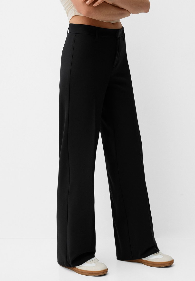 Wide-leg black trousers made of smooth fabric, featuring a zip fly, side pockets, and a tailored fit with a structured look.