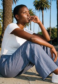 Woman with short hair in white shirt and blue striped pants sits on pavement, resting chin on hand, with palm trees and clear sky behind.