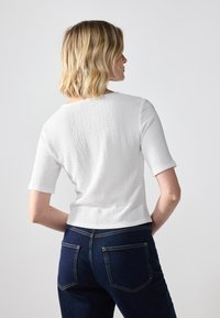 Haut texturé blanc à manches courtes, avec un col rond, associé à un jean bleu foncé affichant des poches arrière et des détails de couture.