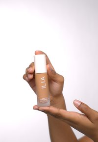 ILIA True Skin Serum Foundation i en gjennomsiktig glassflaske med en beige nyanse, med et hvitt lokk og merket front i fete bokstaver.