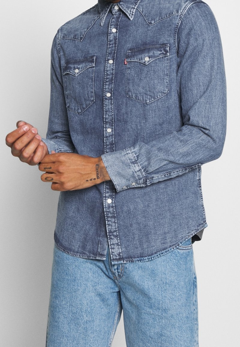 Homme ajustant la manche d'une chemise en denim bleu délavé à boutons, portant un jean denim bleu clair sur fond uni.