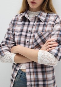 Camicia di flanella a quadri rosa, marrone e blu con bottoni neri; presenta una tasca sul petto e maniche bianche con pizzo su uno sfondo chiaro.