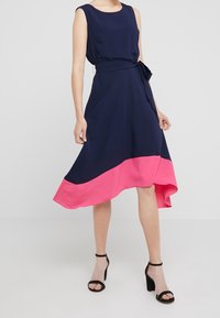 En ärmlös, marinblå klänning med en rosa fåll, tillverkad av lättviktsmaterial. Den har ett knytmidjeband och en asymmetrisk kjol.