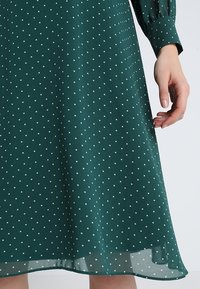 Robe verte à pois avec un ourlet fluide, présentant un tissu doux et un design à manches longues modeste. La robe a des pois blancs uniformément espacés.