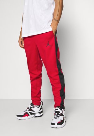 Mężczyzna ubrany w czerwono-czarne spodnie dresowe marki Jordan oraz białe, czarno-czerwone buty sportowe, stojący z jedną ręką w kieszeni.