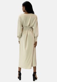 Robe beige à manches longues avec une ceinture à la taille, des plis à l'arrière et une texture lisse. Portée avec des talons noirs avec des brides à la cheville.