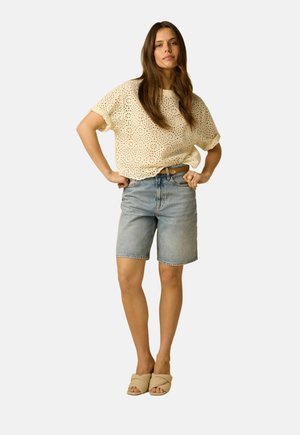 Femme debout, mains sur les hanches, portant un haut crème à œillets, un short en jean longueur genou et des sandales beige à enfiler, sur un fond clair.