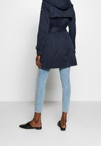 Osoba w granatowym płaszczu, jasnoniebieskich, wzorzystych rurkach i czarnych butach wsuwanych, stojąca na tle gładkiej białej ściany.