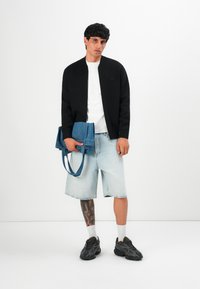 Jeune homme portant une veste noire, un t-shirt blanc, un short en denim clair, des baskets noires, tenant un sac fourre-tout bleu plié, se tenant contre un fond blanc.