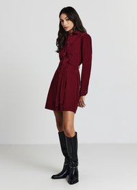 Vestito camicia in velluto a coste bordeaux con cintura in vita, maniche lunghe, bottoni frontali e due tasche sul petto. Indossato con stivali neri al ginocchio.