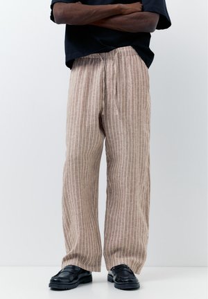 Muž stojaci so založenými rukami, oblečený v béžových ľanových nohaviciach s bielymi vertikálnymi pruhmi a čiernych kožených mokasínach na bielom pozadí.