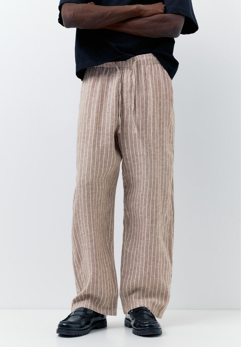 Homme debout, bras croisés, portant un pantalon en lin beige à rayures verticales blanches et des mocassins en cuir noir sur fond blanc.
