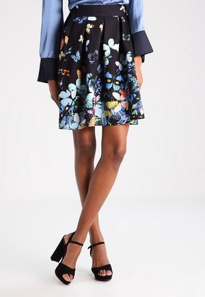 Mujer con una falda negra acampanada con estampados de mariposas de colores, una blusa azul con puños acampanados y sandalias negras de tacón alto.