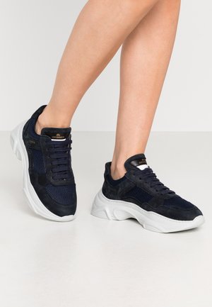 Nogi w granatowych siateczkowych i zamszowych sneakersach z grubymi białymi podeszwami, logo „Copenhagen” widoczne na języku i boku.