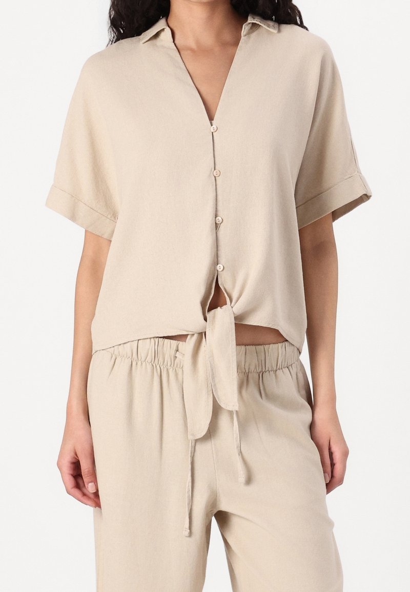 Femme portant une chemise beige à manches courtes boutonnée, nouée à la taille, avec un pantalon beige assorti à taille élastique.
