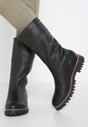 Bottes noires en cuir mi-mollet avec semelles robustes portées sur un pantalon beige ajusté, montrant des détails cousus et de légers plis sur les bottes.