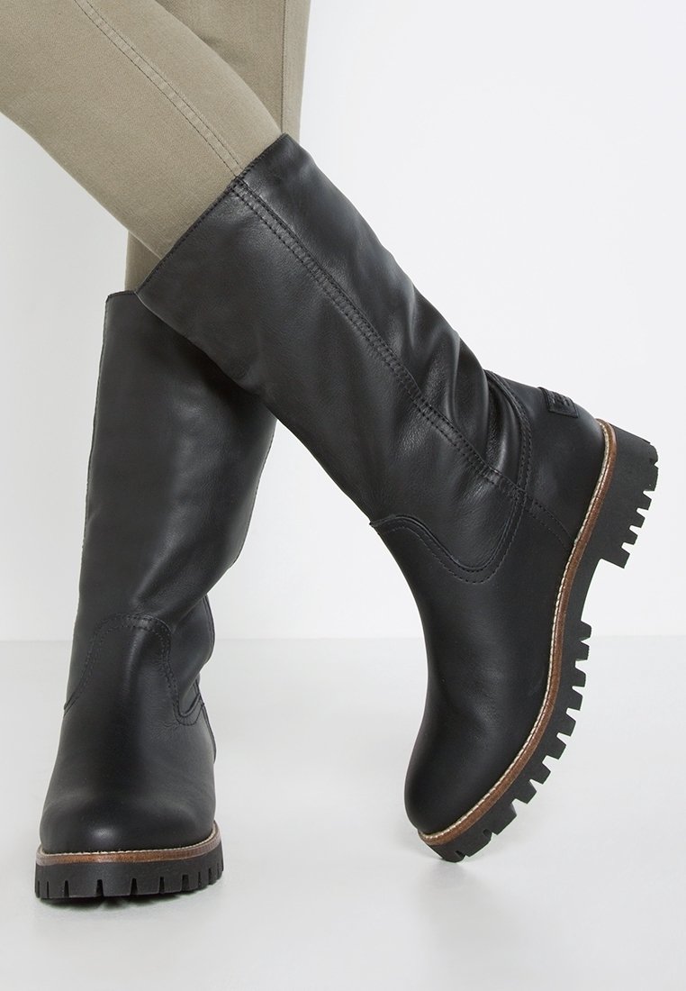 Bottes noires en cuir mi-mollet avec semelles robustes portées sur un pantalon beige ajusté, montrant des détails cousus et de légers plis sur les bottes.