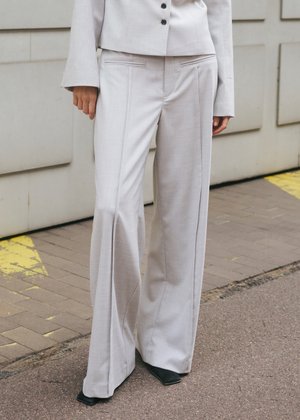 Személy világosszürke, bőszárú nadrágot és hozzá illő blézert visel, valamint fekete hegyes orrú cipőt, egy burkolt felületen áll.