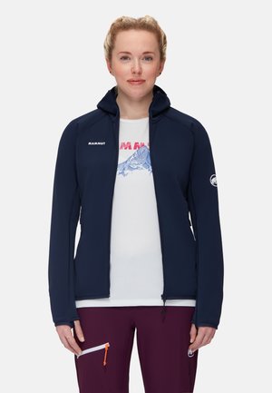 Ung kvinde med lyst hår trukket tilbage iført en marineblå jakke med lynlås, hvid t-shirt med grafik og mørkelilla bukser med lynlåslomme.