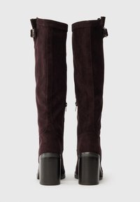 Térdmagasságú barna velúr csizmák, tömbsarokkal, oldalirányú cipzárral és a tetején állítható illeszkedésű csat díszítéssel.