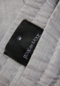 Grå tygetikett med "JUNK de LUXE" i vit text, mot en svart bakgrund, med en liten kronlogotyp ovanför varumärket.