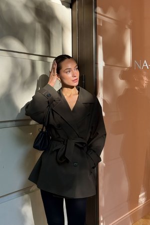 Tumeoliivroheline vööga trench coat laia kraega ja kahe suure nööbiga; koos kitsaste mustade pükste ja väikese musta kotiga.