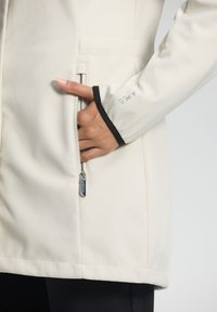 Lichtbeige jasje met een gladde textuur, voorzien van een zijzak met rits en zwarte accenten op de manchetten en onderkant.