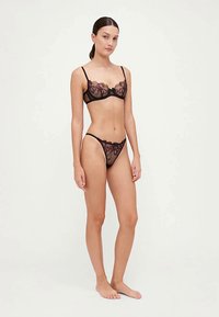 Conjunto de sutiã e fio dental em renda preta com padrões florais intrincados, bordas em forma de ondas e material de malha macia, apresentando alças ajustáveis e um laço delicado.