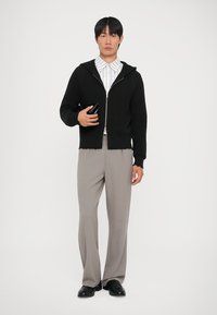 Svart zip-up-tröja med ett texturerat mönster, bärs över en vit randig skjorta, ihop med ljusgrå plisserade byxor och svarta skor.