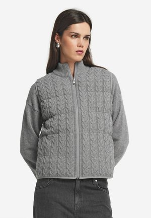 Femme portant un gilet zippé en tricot gris sur un pull à manches longues, associé à un jean noir et des boucles d'oreilles rondes en argent.