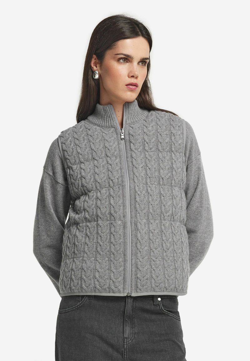 Kvinna iklädd en grå kabelstickad zip-up väst över en långärmad tröja, parat med svarta jeans och silverfärgade runda örhängen.