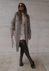 Lungo cappotto grigio con dettagli a bottoni sopra un maglione crema, abbinato a una gonna bianca e stivali alti fino al ginocchio marroni. Gli accessori includono occhiali da sole.