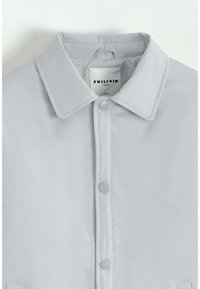 Chemise gris clair avec un col, fermeture à boutons-pression et une étiquette "FRILIVIN" au niveau du col. Tissu lisse avec une finition mate.