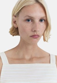 Top de color claro a rayas con tirantes anchos. La modelo tiene el pelo corto y ondulado de color rubio y una expresión neutral. Su piel tiene pecas visibles.