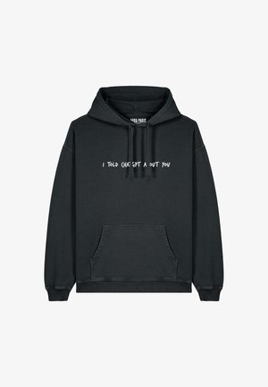 Haut à capuche oversized noir avec le texte blanc "JE L'AI DIT À CH TGBT À PROPOS DE TOI," associé à un pantalon cargo beige et des baskets blanches.