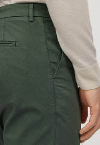 Gröna bomullsshorts med en slät textur, med en sidoficka och en bakficka med en dold söm. Handen vilar på höfterna.