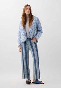 Jachetă albastră matlasată cu cusături în formă de diamant, asociată cu pantaloni din denim în dungi, cu talie înaltă, în nuanțe de albastru și sandale albastre cu accesorii metalice.