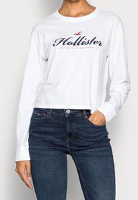 Vit långärmad T-shirt med "Hollister Southern California"-logotyp i marinblått och röd, kombinerad med mörkblå jeans. Materialet verkar vara mjuk bomull.