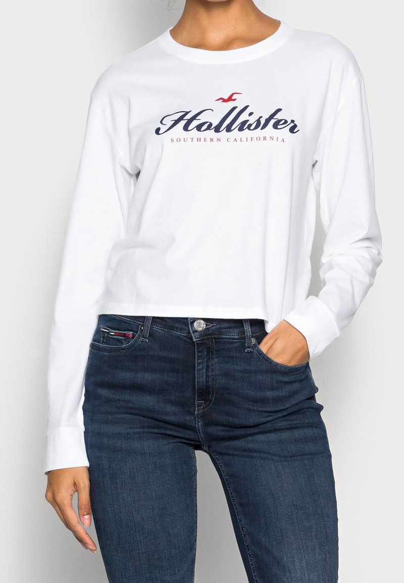 Vit långärmad T-shirt med "Hollister Southern California"-logotyp i marinblått och röd, kombinerad med mörkblå jeans. Materialet verkar vara mjuk bomull.