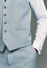 Gilet texturé bleu clair avec cinq boutons, une poche passepoilée, associé à une chemise blanche avec des boutons de poignet noirs.