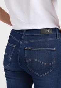 Dunne blauwe jeans van denim met een gladde textuur. Voorzien van oranje stiksels langs de gebogen zakken en een zwart Lee merklabel.