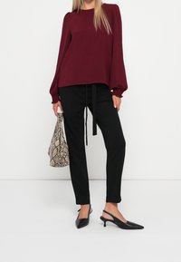 Burgunderfarbener, strukturierter Langarmoberteil, schwarze, schmal zulaufende Hose mit Bindegürtel, schwarze Slingback-Absätze und eine beige Handtasche aus Schlangenleder.