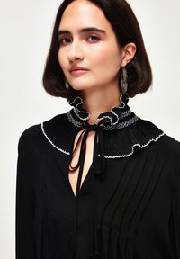 Schwarze Bluse mit einem Rüschensaum in Weiß, Knopfleiste mit Bändchen und vertikalen Falten, aus einem glatten Stoff hergestellt.