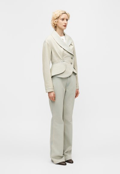 Νεαρή γυναίκα με κοντά σγουρά ξανθά μαλλιά, που φοράει ένα ανοιχτό μπεζ, ραμμένο στα μέτρα, διπλόμπουτο σακάκι και ταιριαστό παντελόνι φαρδιάς γραμμής.