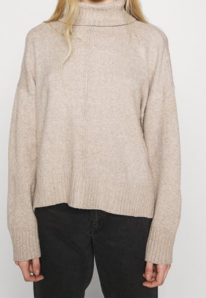 Personne portant un pull beige en maille col roulé à manches longues et un pantalon noir, montré des épaules jusqu'aux hanches.