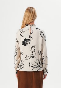 Donna con lunghi capelli biondi che indossa una blusa beige con stampato floreale nero e colletto arricciato, abbinata a una gonna marrone, voltata di spalle.